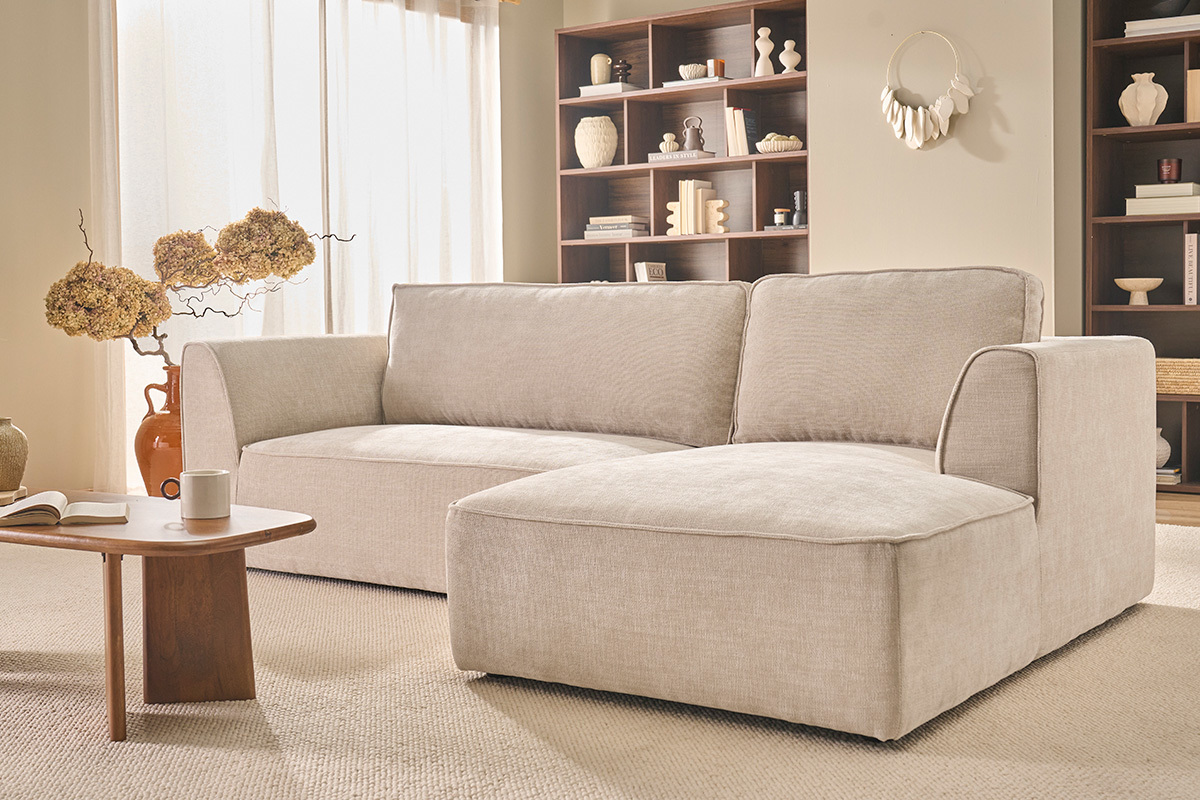 Canap� d'angle droit beige 3 places en tissu velours, vue de 3/4, salon avec �tag�res au fond.