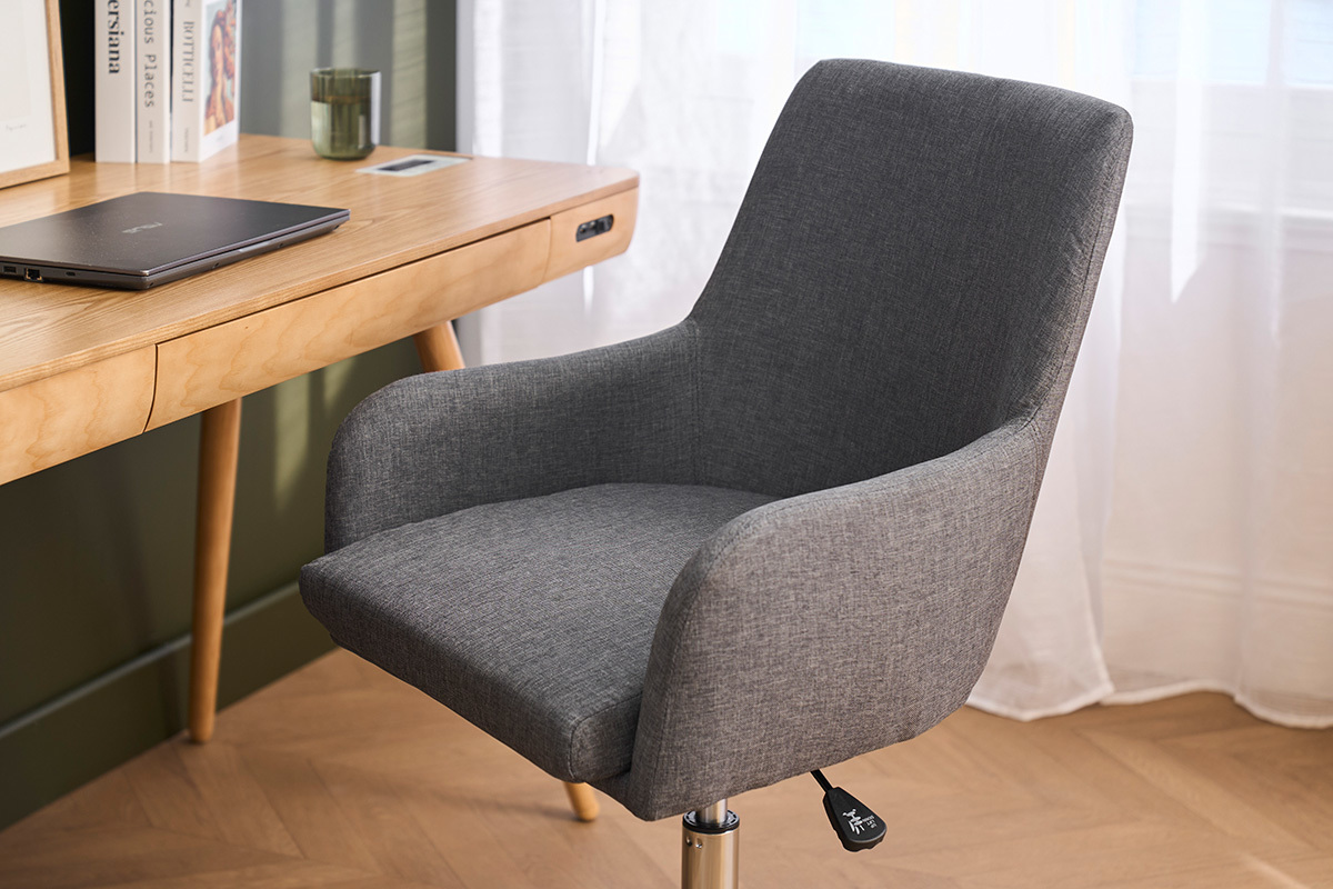 Chaise de bureau en tissu gris, vue de 3/4, avec accoudoirs devant un bureau en bois.