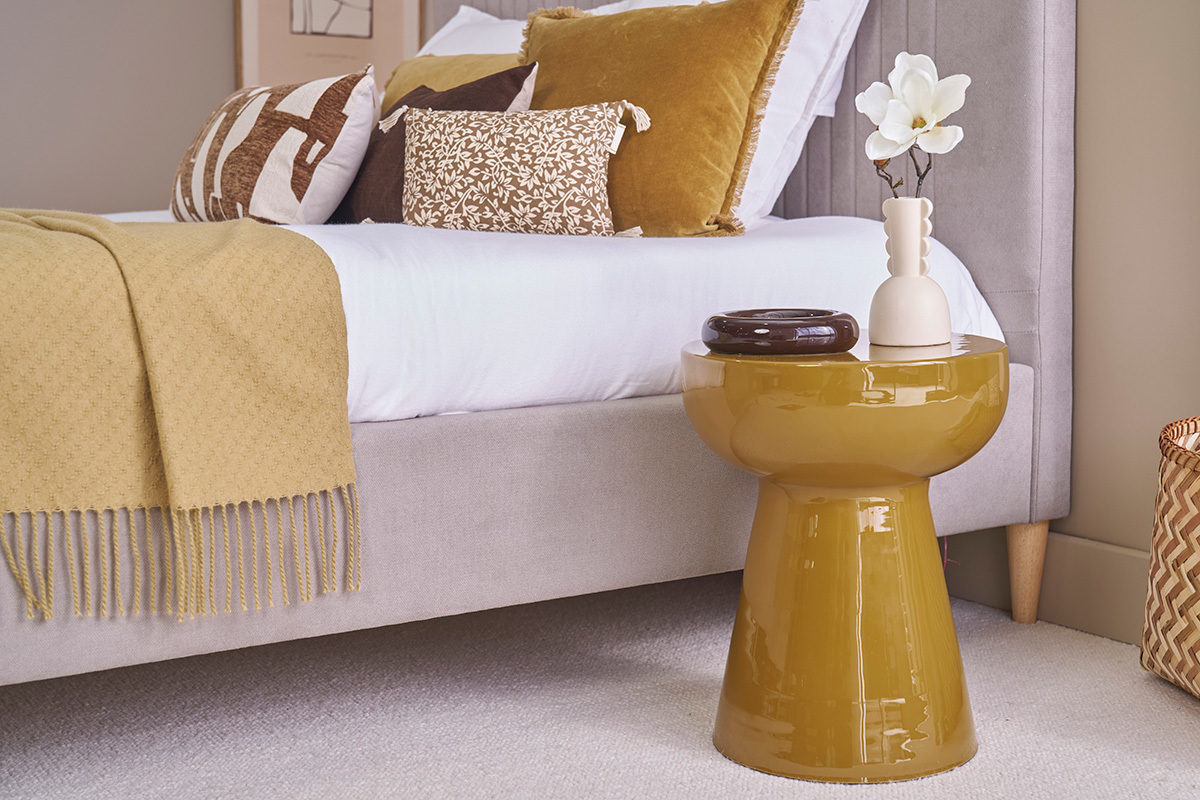 Table d'appoint ronde en m�tal �maill� bronze, pr�s d'un lit avec coussins d�coratifs.